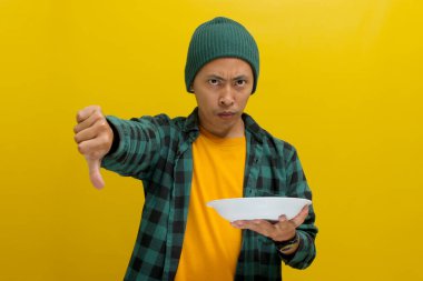 Hoşnut olmayan Asyalı genç bir adam, bereli bir şapka ve gündelik bir gömlek giyer, bir başparmak aşağı vererek memnuniyetsizlik ifade eder, yiyecek için olumsuz bir eleştiri, boş bir beyaz tabak tutarken
