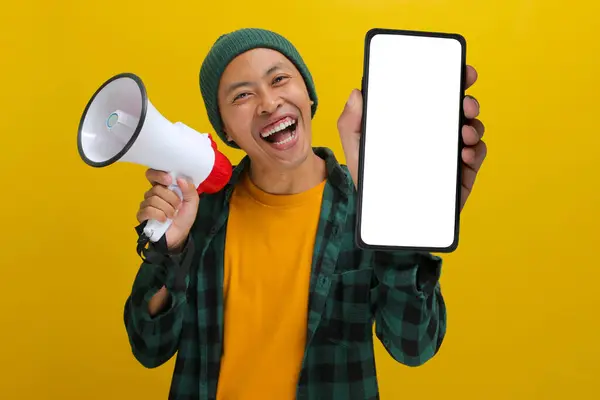 Heyecanlı Asyalı adam elinde indirimli satış, özel teklif ya da yeni bir uygulamayı tanıtmak için elinde megafon tutarken boş ekranlı bir telefon gösteriyor. Sarı arka planda izole edilmiş.