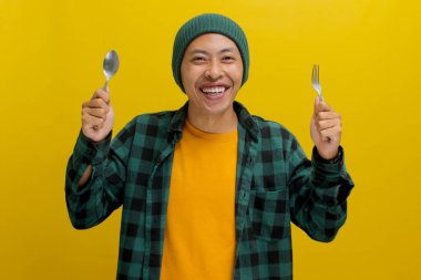 Şapkalı ve rahat gömlekli neşeli Asyalı bir adam elinde çatal ve kaşıkla kameraya bakıp heyecanla gülümserken lezzetli bir yemek bekliyor.