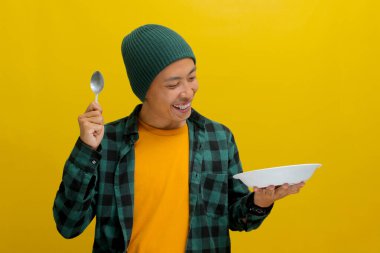 Asyalı heyecanlı genç bir adam, beresi ve günlük gömleği olan, elinde boş bir tabağa bakarken bir kaşık tutuyor, sarı bir arka planda izole edilmiş.