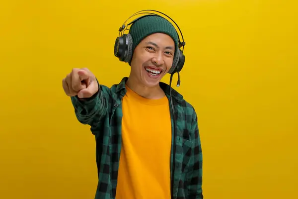 stock image Excited Asian man in a beanie and casual clothes, wearing headphones, points excitedly towards the camera after noticing something amazing while listening to music. Isolated on a yellow background.