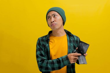 İşsiz Asyalı bir adam boş cüzdanına bakarken şaşkın ve suskun görünüyor, borçtan kaynaklanan maddi sıkıntılarla karşı karşıya, geliri, maaşı ya da maaşı yok, sarı bir arka planda izole edilmiş.