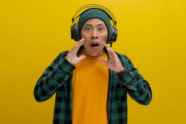 Asian man in a beanie and casual clothes, wearing headphones, looks shocked with his mouth agape after hearing something surprising on his music. Isolated on a yellow background. clipart