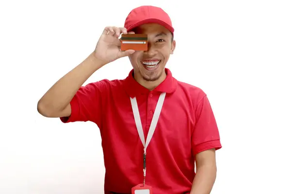 stock image An excited Asian delivery man or courier playfully covers half of his face with a credit card, isolated on a white background. Concepts of online transactions, secure payments, customer satisfaction