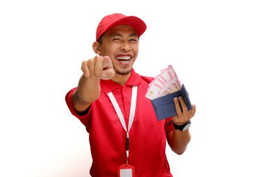 Asian delivery man or courier points towards the camera while holding money banknotes, isolated on white background. Concepts of professionalism, customer engagement, and secure monetary transactions. clipart