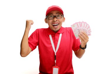 Excited Asian delivery man or courier raises his fist in triumphant YES gesture while holding money banknotes in his hand, isolated on white background. Concepts of victory, financial gains, triumphs clipart