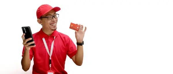 stock image Asian delivery man or courier holds a smartphone in one hand and a credit card in the other, isolated on a white background. Concepts of mobile payments, e-commerce deliveries, and online shopping