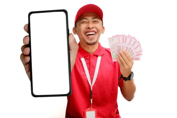 Stock image Asian delivery man or courier Showing Blank Phone Screen towards the camera, while holding Money Banknotes, Isolated on white background, new apps, online shopping, logistics, and financial management