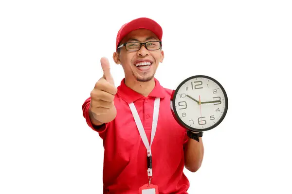 stock image Excited Asian delivery man or courier smiles and shows a thumbs-up gesture to the camera while holding a clock in his hand, isolated on a white background