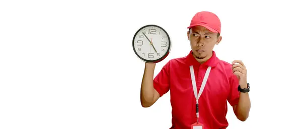 stock image Confused Asian delivery man or courier holds a wall clock while looking at an empty copy space, isolated on white background. Concept of time management, deadlines, or scheduling in delivery services