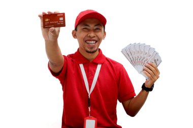 Kırmızı polo tişört giyen Asyalı teslimatçı beyaz arka planda kredi kartı ve dolar banknotları tutuyor. Teslimat hizmetleri, finansal işlemler, e-ticaret ve müşteri memnuniyeti.