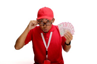 Asyalı kurye beyaz arka planda Endonezya parası tutarken şok ve heyecan ifade ediyor. Mali sürpriz kavramı, e-ticaret başarısı, teslimat hizmetleri ve olumlu tepkiler.