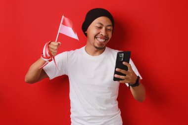 Heyecanlı Endonezyalı genç adam, Endonezya Bağımsızlık Günü 'nü 17 Ağustos' ta kutlarken elinde kırmızı arka planda izole edilmiş bir şekilde Endonezya bayrağı ve telefonu tutuyor..