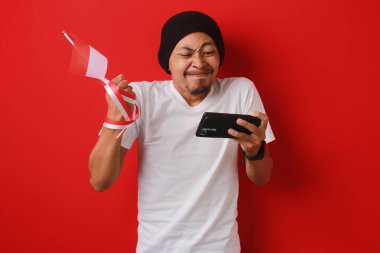 Sinirli Endonezyalı genç adam Endonezya Milli Takımı maçını 17 Ağustos 'ta Endonezya Bağımsızlık Günü' nde kırmızı arka planda izole edilmiş Endonezya bayrağını tutarken telefonundan izliyor.