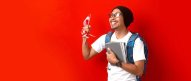 Endonezyalı erkek öğrenci Endonezya Bağımsızlık Günü 'nü 17 Ağustos' ta kutluyor. Elinde Endonezya bayrağı, bir sırt çantası ve kırmızı arka planda izole edilmiş bir kitapla.