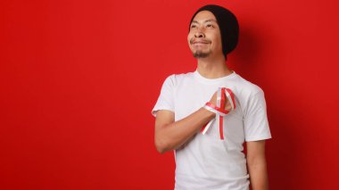 Beyaz tişörtlü ve bereli mutlu Endonezyalı adam Endonezya milli marşını dinlerken elini göğsüne koydu. Kırmızı arkaplanda izole edilmiş.