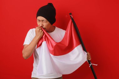 Beyaz gömlekli ve bereli Endonezyalı genç adam Endonezya bayrağını öpüyor ve 17 Ağustos 'ta Bağımsızlık Günü' nü kutluyor. Sahne kırmızı arka planda izole edilmiştir..