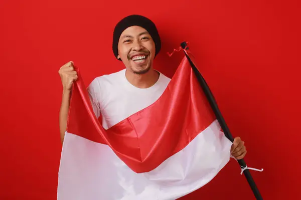 stock image Happy and excited Indonesian man holds Indonesia's flag to celebrate Indonesia Independence Day. Isolated on a red background.