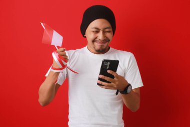 Heyecanlı Endonezyalı genç adam, Endonezya Bağımsızlık Günü 'nü 17 Ağustos' ta kutlarken elinde kırmızı arka planda izole edilmiş bir şekilde Endonezya bayrağı ve telefonu tutuyor..