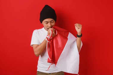 Beyaz gömlekli ve bereli Endonezyalı genç adam Endonezya bayrağını öpüyor ve 17 Ağustos 'ta Bağımsızlık Günü' nü kutluyor. Sahne kırmızı arka planda izole edilmiştir..