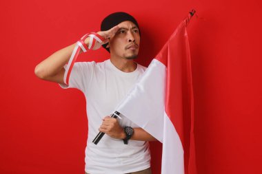 Endonezyalı genç adam, Endonezya bayrağı taşıyan beyaz bir gömlek ve bere giyerek 17 Ağustos 'ta Bağımsızlık Günü' nü kutlarken selam veriyor. Kırmızı arkaplanda izole edilmiş.