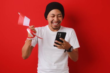 Heyecanlı Endonezyalı genç adam, Endonezya Bağımsızlık Günü 'nü 17 Ağustos' ta kutlarken elinde kırmızı arka planda izole edilmiş bir şekilde Endonezya bayrağı ve telefonu tutuyor..