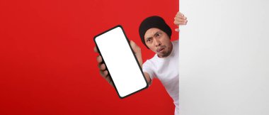 Şok edici Endonezyalı adam beyaz bir duvarın arkasından kameraya boş beyaz bir ekranla telefon gösteriyor, yeni uygulamalar veya Endonezya Bağımsızlık Günü 'nde beklenmedik özel bir teklif sunuyor. Kırmızı Arkaplan