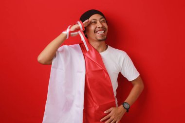 Endonezyalı genç adam, Endonezya bayrağı taşıyan beyaz bir gömlek ve bere giyerek 17 Ağustos 'ta Bağımsızlık Günü' nü kutlarken selam veriyor. Kırmızı arkaplanda izole edilmiş.