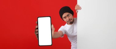 Şok edici Endonezyalı adam beyaz bir duvarın arkasından kameraya boş beyaz bir ekranla telefon gösteriyor, yeni uygulamalar veya Endonezya Bağımsızlık Günü 'nde beklenmedik özel bir teklif sunuyor. Kırmızı Arkaplan
