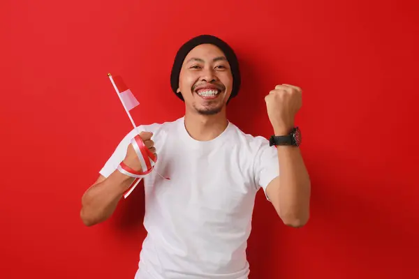 stock image Excited Indonesian man holds the Indonesian flag and raises his fist in celebration of Indonesian Independence Day on August 17, isolated on a red background