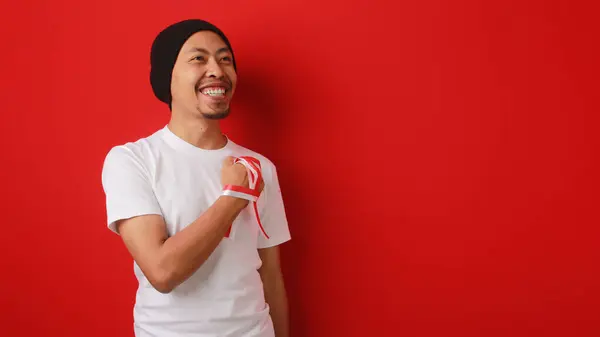 stock image Happy Indonesian man in a white T-shirt and beanie hat, with an Indonesia flag ribbon, puts his hand on his chest while listening to the Indonesia national anthem. Isolated on a red background.