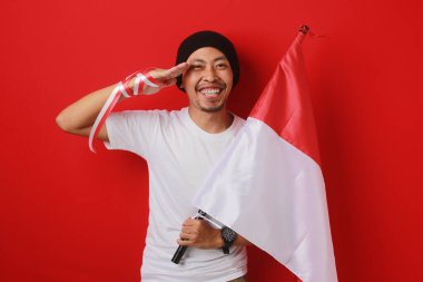 Endonezyalı genç adam, Endonezya bayrağı taşıyan beyaz bir gömlek ve bere giyerek 17 Ağustos 'ta Bağımsızlık Günü' nü kutlarken selam veriyor. Kırmızı arkaplanda izole edilmiş.