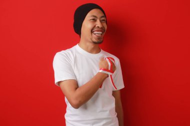 Beyaz tişörtlü ve bereli mutlu Endonezyalı adam Endonezya milli marşını dinlerken elini göğsüne koydu. Kırmızı arkaplanda izole edilmiş.