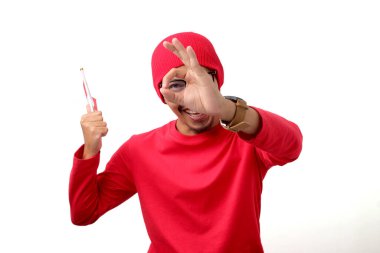 Excited Indonesian youth peeks through an OK gesture while looking at the camera and holding the Indonesian flag during Indonesia Independence Day celebration on August 17 isolated on white background clipart