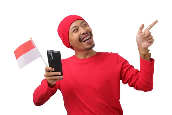 stock image Indonesian man in a red long sleeve shirt points at empty copy space while holding the Indonesian flag and phone during Indonesia Independence Day Celebration on August 17 isolated on white background