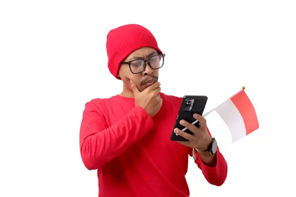 stock image Confused young Indonesian man in a red shirt looks at his phone thoughtfully while holding the Indonesian flag during Indonesia Independence Day celebration on August 17, isolated on white background