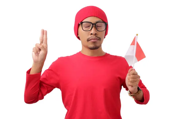 stock image Indonesian man wearing red shirt and a beanie makes a promises gesture while holding the Indonesian flag during Indonesian Independence Day celebration on August 17, isolated on a white background.