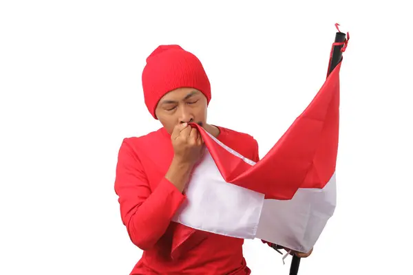 stock image Young Indonesian man in a red long sleeve shirt and beanie is kissing the Indonesian flag, celebrating Indonesia's Independence Day on 17 August. The scene is isolated on a white background.