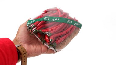 A hand holds a package of fresh red chili peppers, wrapped in plastic and secured with a green 