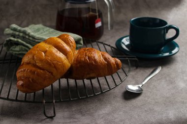 Soğutma rafında taze pişmiş kruvasan, yanında bir fincan çay ve çaydanlık. Kahvaltı ya da brunch konsepti