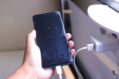 Trendeki elektrik prizinden şarj olan akıllı telefonu tutan bir adamın yakın plan fotoğrafı. Telefonun ekranı siyah ve şarj kablosu telefona ve prize bağlı.