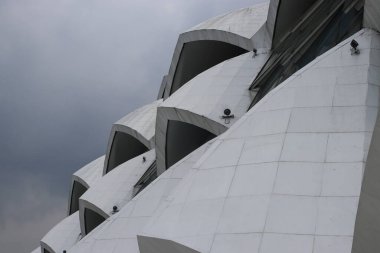 Modern bir binanın benzersiz çatı çizgisine yakın çekim. Tekrarlayan kıvrımlı ve açılı bölümler. Beyaz cephe arka plandaki bulutlu gökyüzü ile çelişiyor