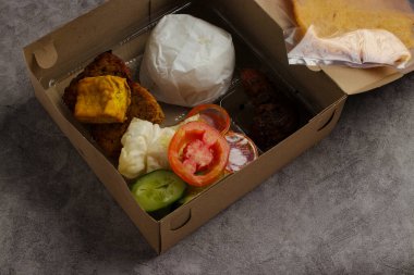 Nasi Kotak featuring ayam bakar (roasted chicken), served with a mound of white rice, a crispy cracker, tempeh, tofu, sambal, and fresh vegetables like cucumber and tomato. clipart