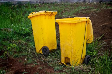 Two yellow recycling bins sitting on a grassy patch of ground, with some dirt and weeds visible. One bin is closed with the lid shut, while the other has its lid open clipart