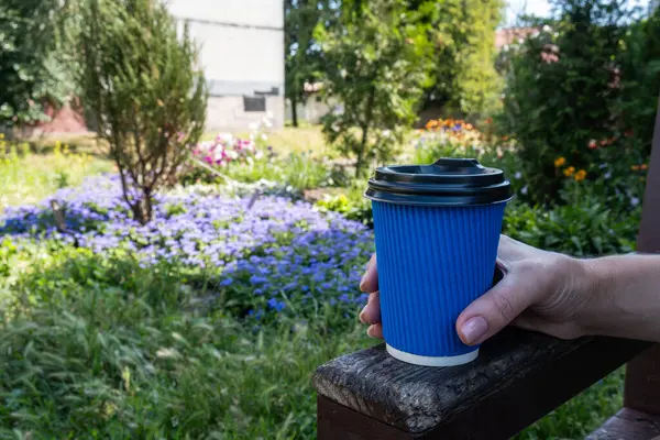 Blue paper coffee cup in the hand of a girl close-up against the background of a city flower bed with flowers. High quality photo
