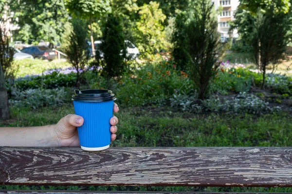 A blue coffee paper cup in the girl\'s hand, placed on the wooden back of a street bench. High quality photo