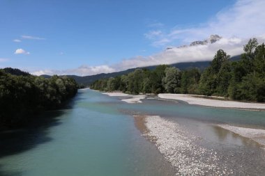 Lech Nehri 'nin turkuaz suyu daimi akışında dağlarla ve uzaktaki beyaz bulutlarla geniş nehir yatağında akıyor.