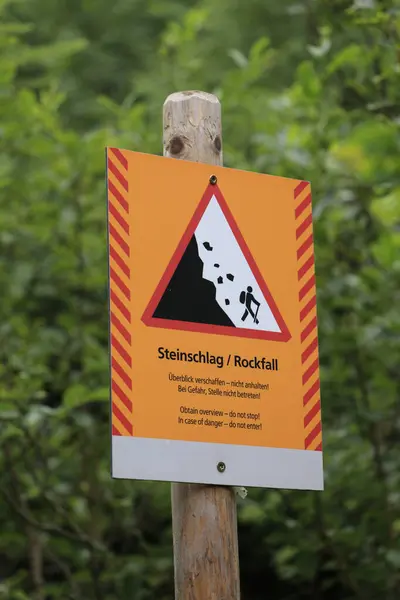 bilingual hazard sign on a hiking trail warning for rock fall. English translation below German text.