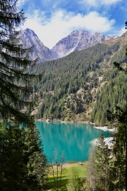 Antholz Gölü 'nün zümrüt yeşili suyu ve etrafındaki ormanın kozalaklı ağaçlarının arasından görünen yüksek dağlar. 