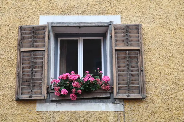 old window in a stone wall with wooden shutters and geraniums on the window sill